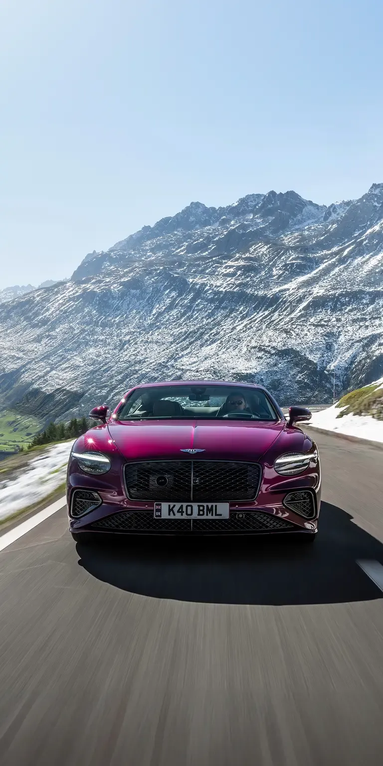 Portrait shot of a magenta Bentley Continental GT approaching on an alpine pass with snowbanks and rugged mountains behind.